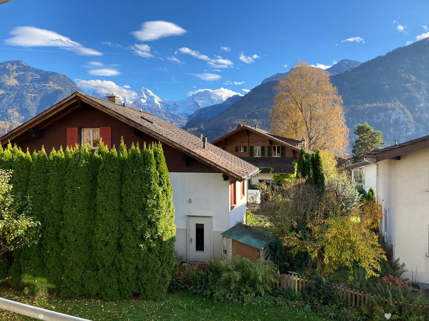 A two-story house with a brown roof and white walls, surrounded by lush greenery and trees, with a scenic mountain view in the background.