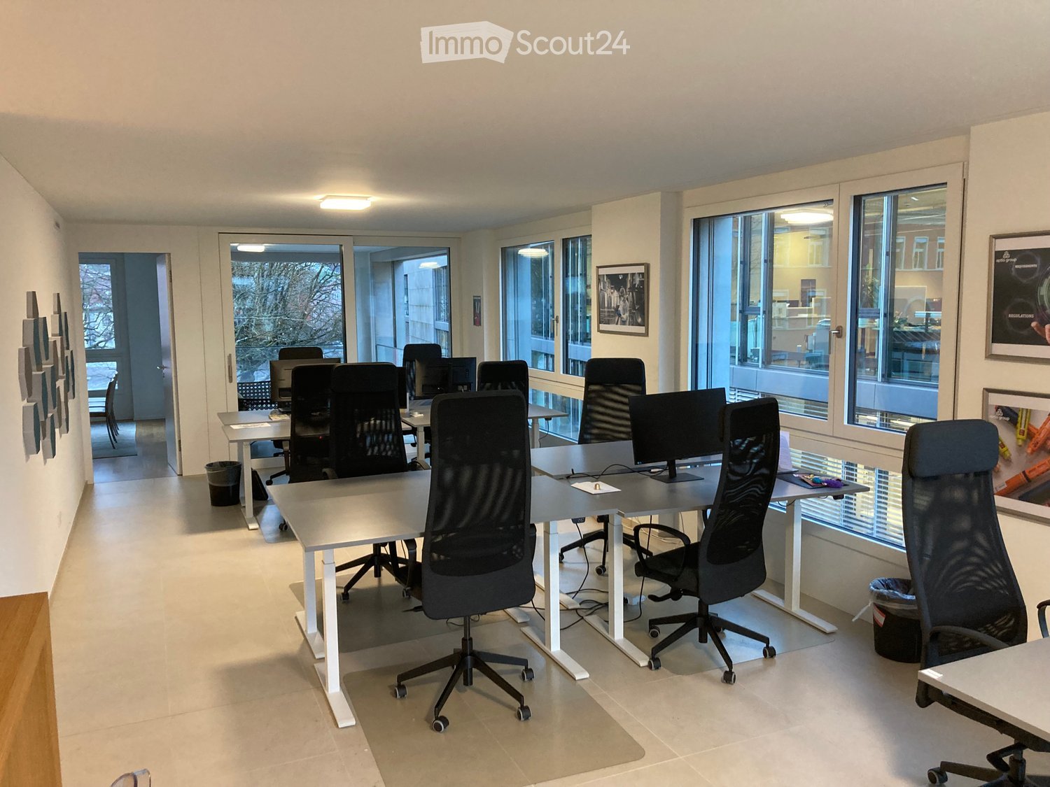 Office room with desks and chairs, large windows, white walls, ceiling lights, black framed prints on the wall, waste bins, trash cans.
