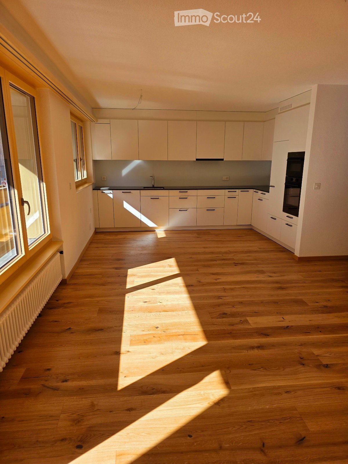 Empty kitchen with white cabinets, wooden floor, and large window allowing natural light to enter.