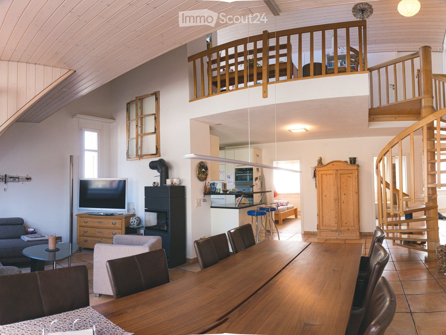 Open-plan living and dining area with a fireplace, television, sofa, coffee table, dining table with chairs, and a kitchen area with stools.