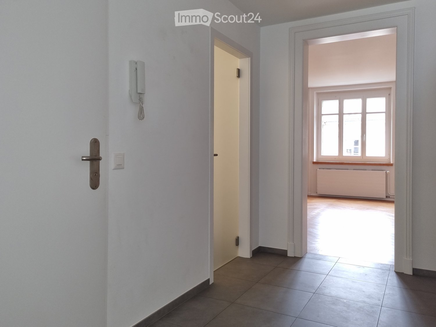 Empty hallway with tiled floor, white walls, and two doors leading to other rooms. One door has a visible telephone and light switch.