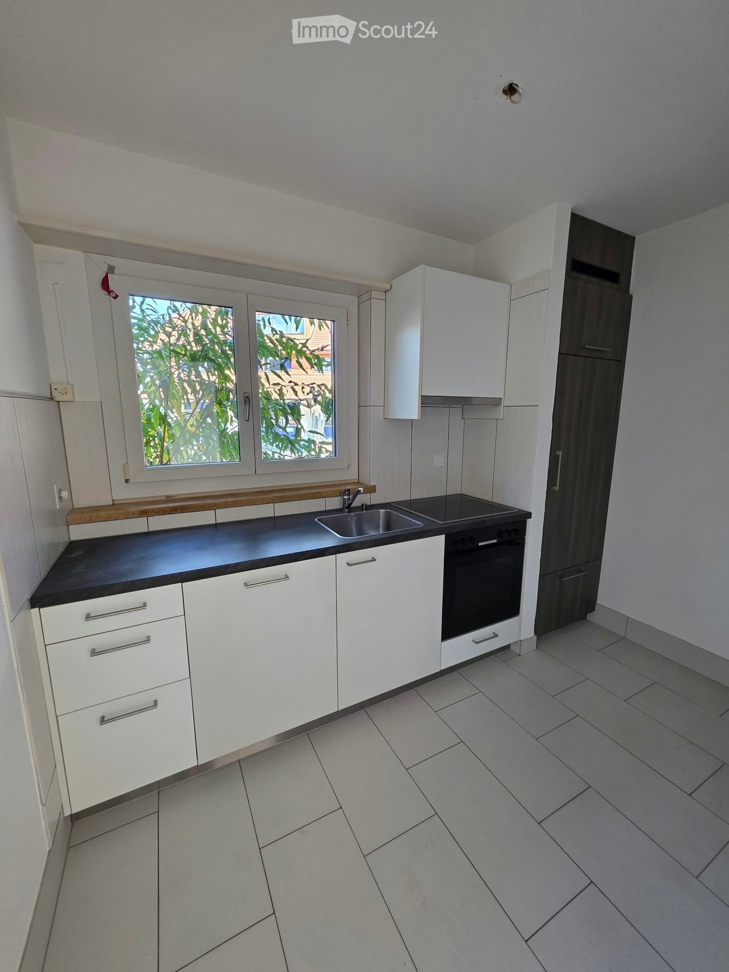 Modern kitchen with white cabinets, black countertop, stainless steel sink, and built-in oven. Features tiled flooring and large windows with a view of greenery.