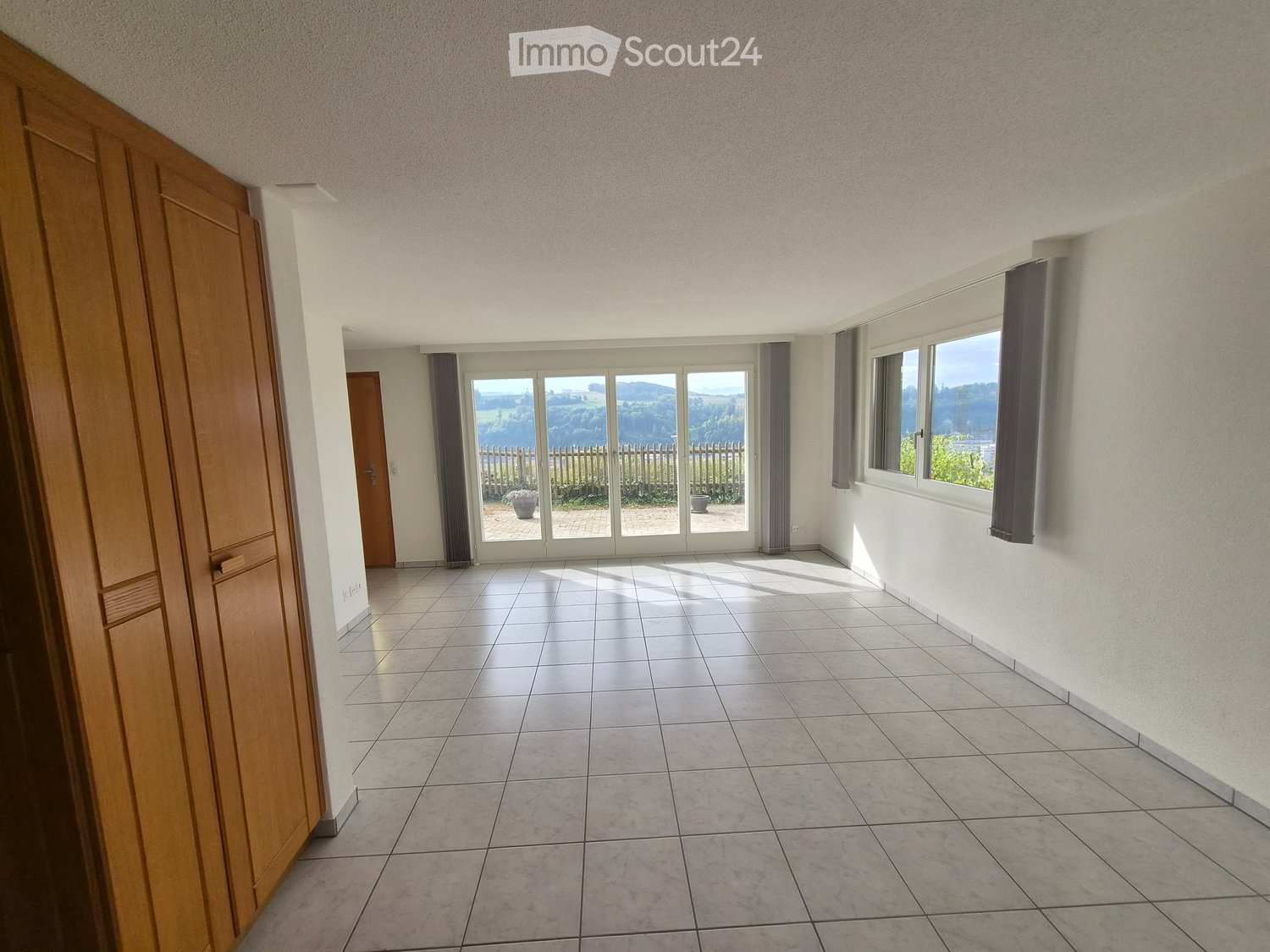 Empty hallway with tiled floor, white walls, large windows, and sliding glass doors leading to an outdoor area.