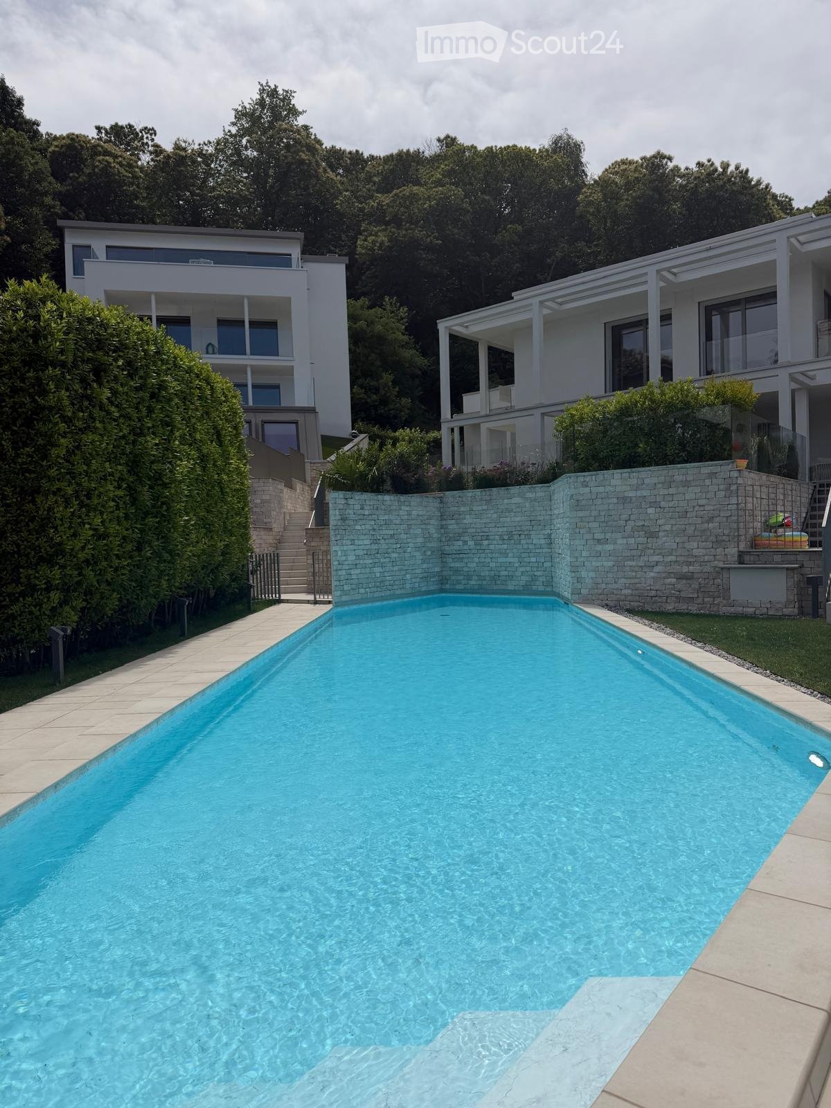 Modern outdoor area with a large rectangular swimming pool surrounded by a stone wall, steps leading up to a modern house with large windows and balconies, lush greenery, and a staircase leading to the house.