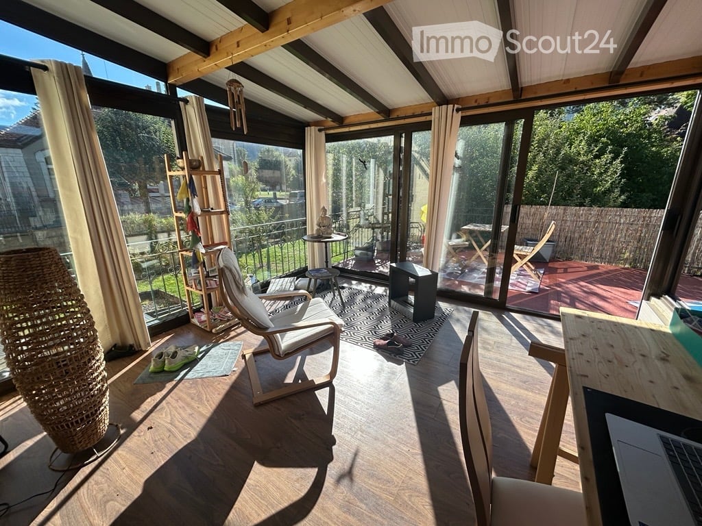 Bright, airy room with wooden flooring, large windows, and a balcony. Features a white chair, small table, stool, laptop, and decorative items.