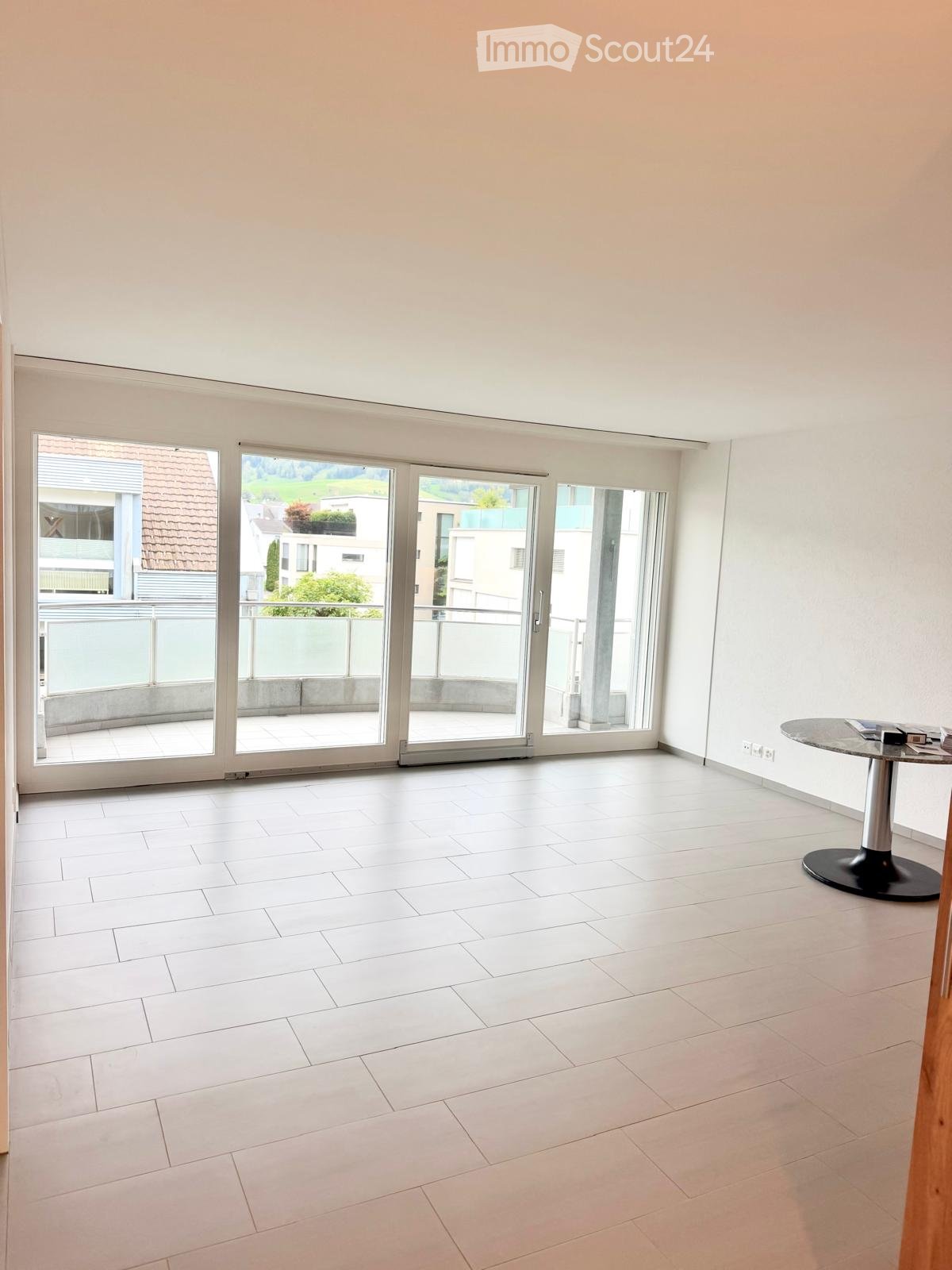 Empty room with white tiled flooring, large sliding glass doors leading to a balcony, and a small round table with books in the corner.