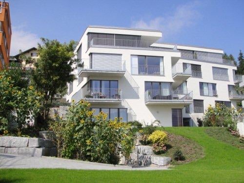 Modern white multi-story apartment building with balconies and large windows, surrounded by a well-maintained garden with sunflowers and shrubs.