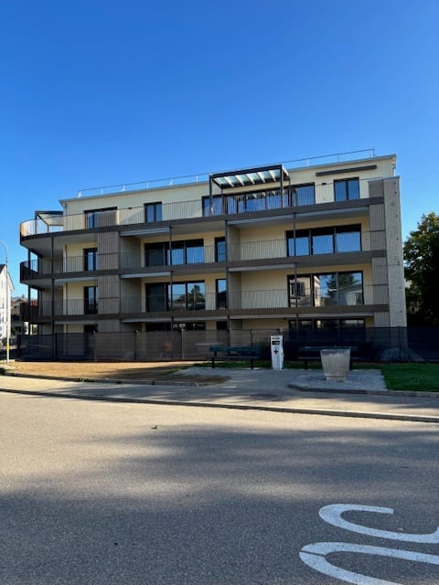 Modern multi-story apartment building with balconies and large windows, surrounded by a small landscaped area with benches.