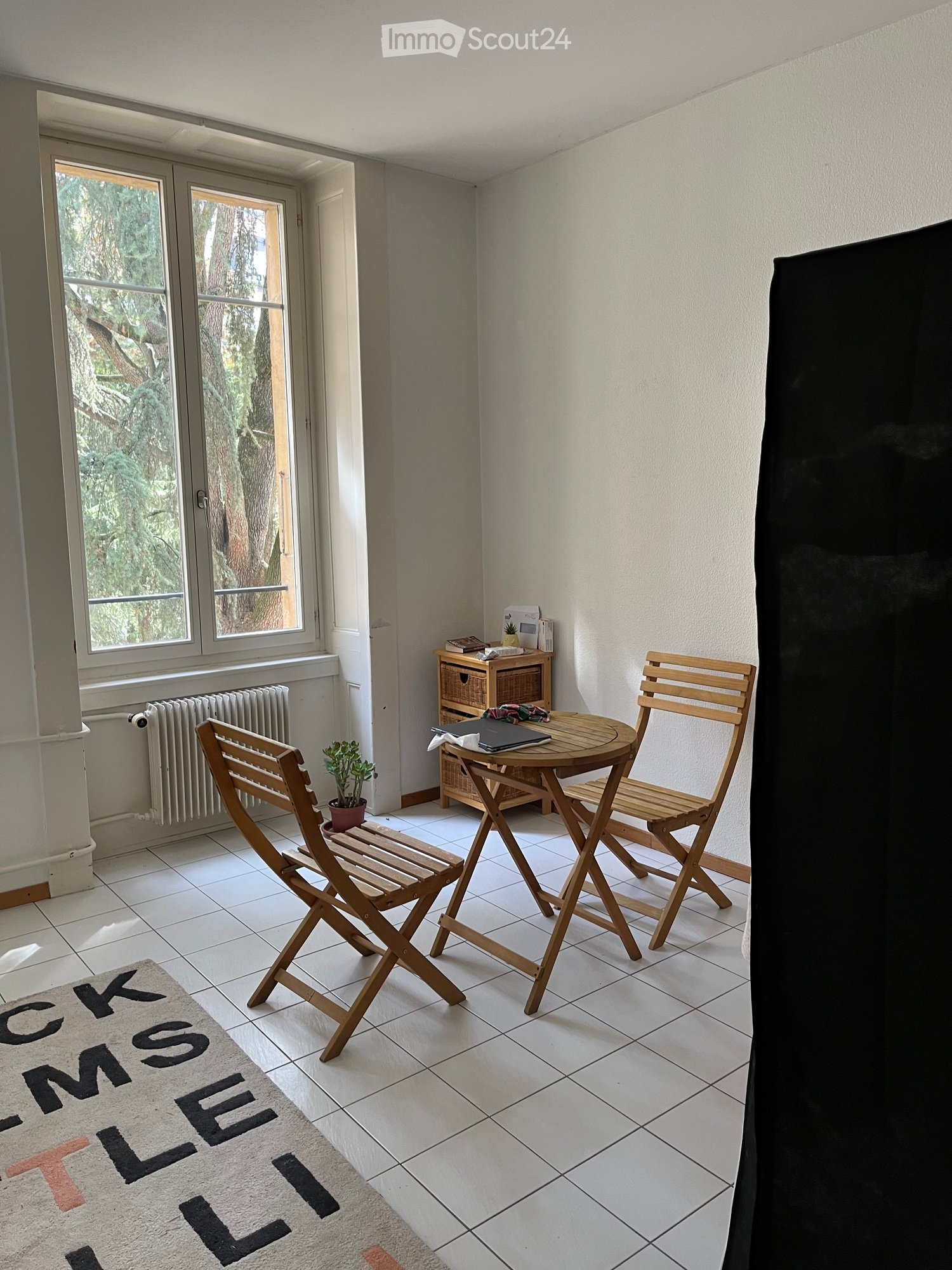 Balcony with white tiled floor, wooden table and chairs, small cabinet, potted plant, and a view of greenery outside.