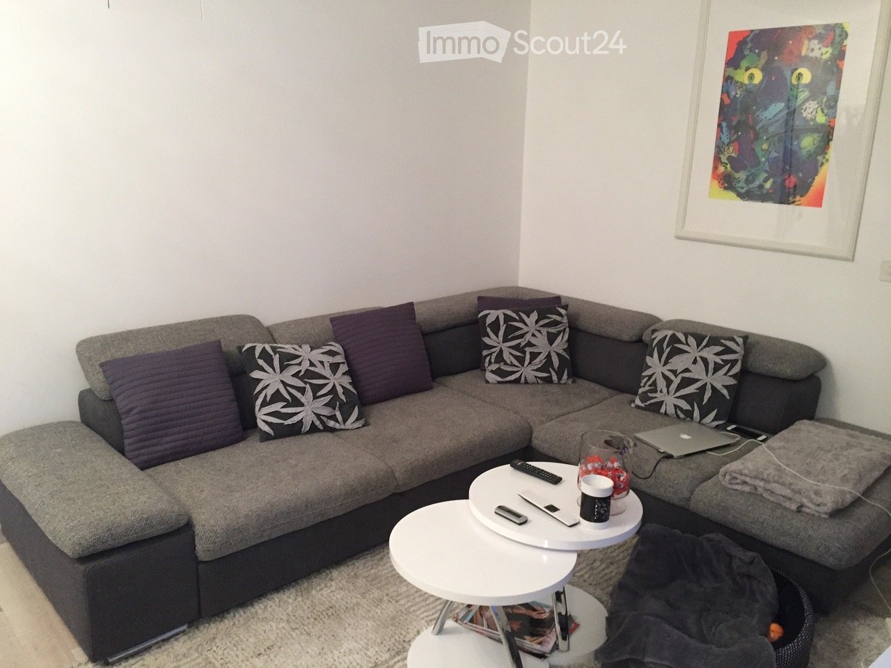 Modern living room with a grey sectional sofa, multiple throw pillows, two white coffee tables, a laptop, and a cup on the coffee table.