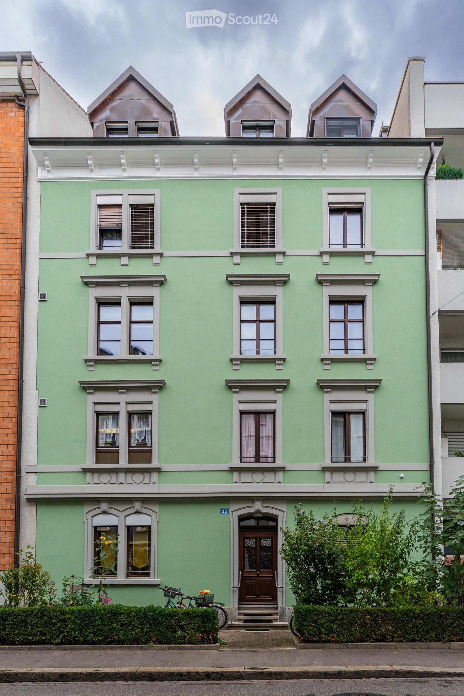 Three-story green house with white trim, multiple windows, decorative roofline, and a bicycle parked near the entrance.