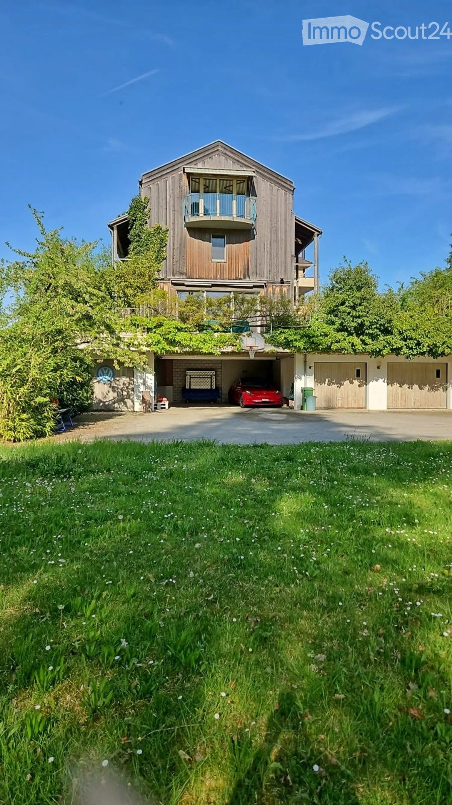 A two-story wooden house with a balcony, garage, and a basketball hoop. The house is surrounded by greenery and a well-maintained lawn.