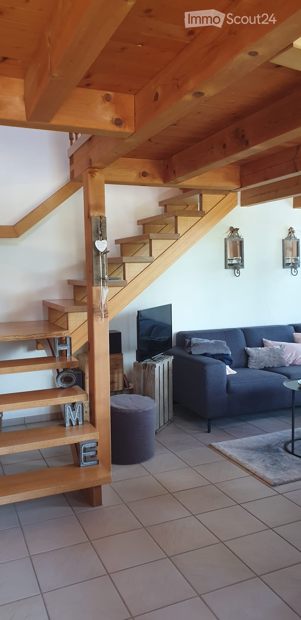 Living room with a wooden staircase leading to an upper level, a grey sofa with cushions, a round grey stool, a small round table, and a television on a wooden stand.