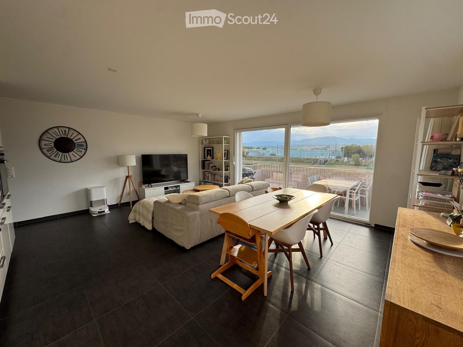 Modern living and dining area with a sofa, dining table, chairs, television, shelves, and a large window offering a scenic view.