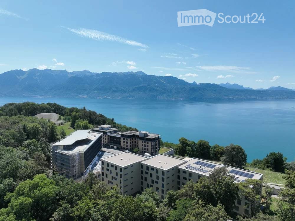 Aerial view of a modern building complex surrounded by trees, overlooking a lake and mountain range.