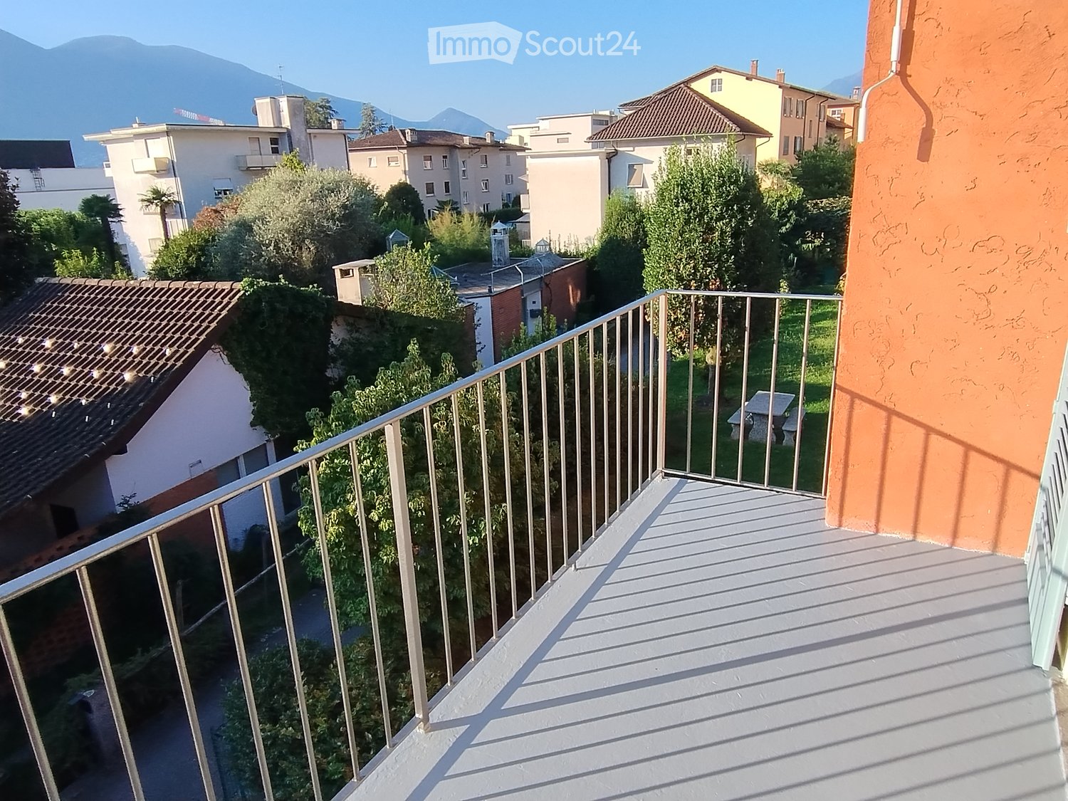 Balcony with metal railing, overlooking a residential area with houses, trees, and a clear blue sky.
