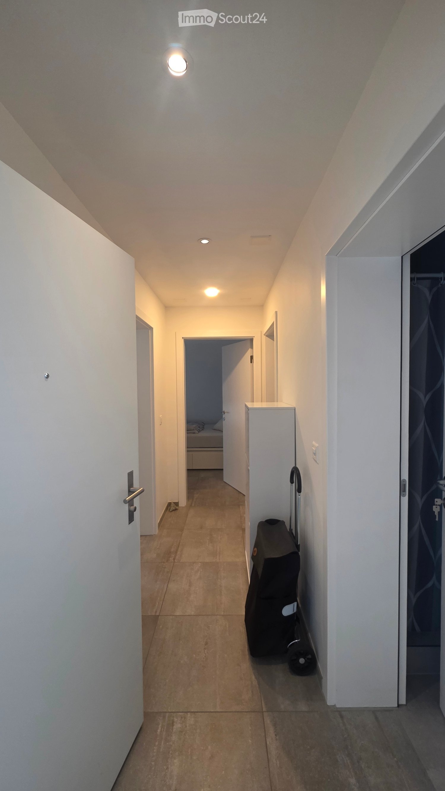 A narrow hallway with beige tiled flooring, white walls, and recessed lighting. A suitcase is placed against the wall on the right side, and a door is visible at the end of the hallway.