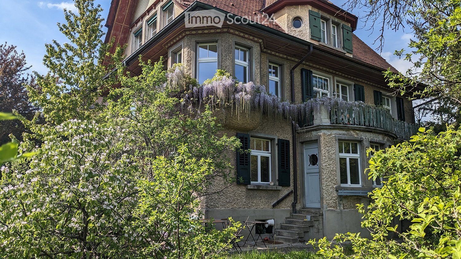 A two-story house with a stone facade, multiple windows with shutters, and a balcony adorned with flowering plants. The entrance features a small staircase leading to a blue door, surrounded by lush greenery and trees.