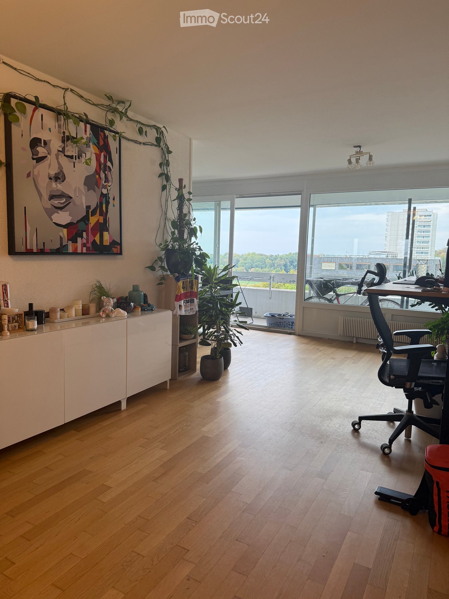 Bright room with wooden flooring, large windows offering a city view, a desk with a chair, a cabinet with decorative items, and several potted plants.