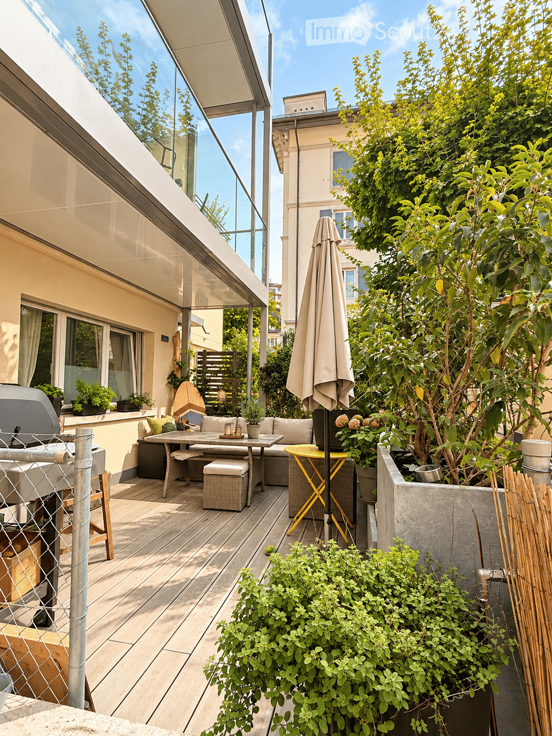 A modern rooftop terrace with wooden decking, a dining table with chairs, a sofa, potted plants, and an umbrella. The area is enclosed by a glass railing and features a grill on the left side.