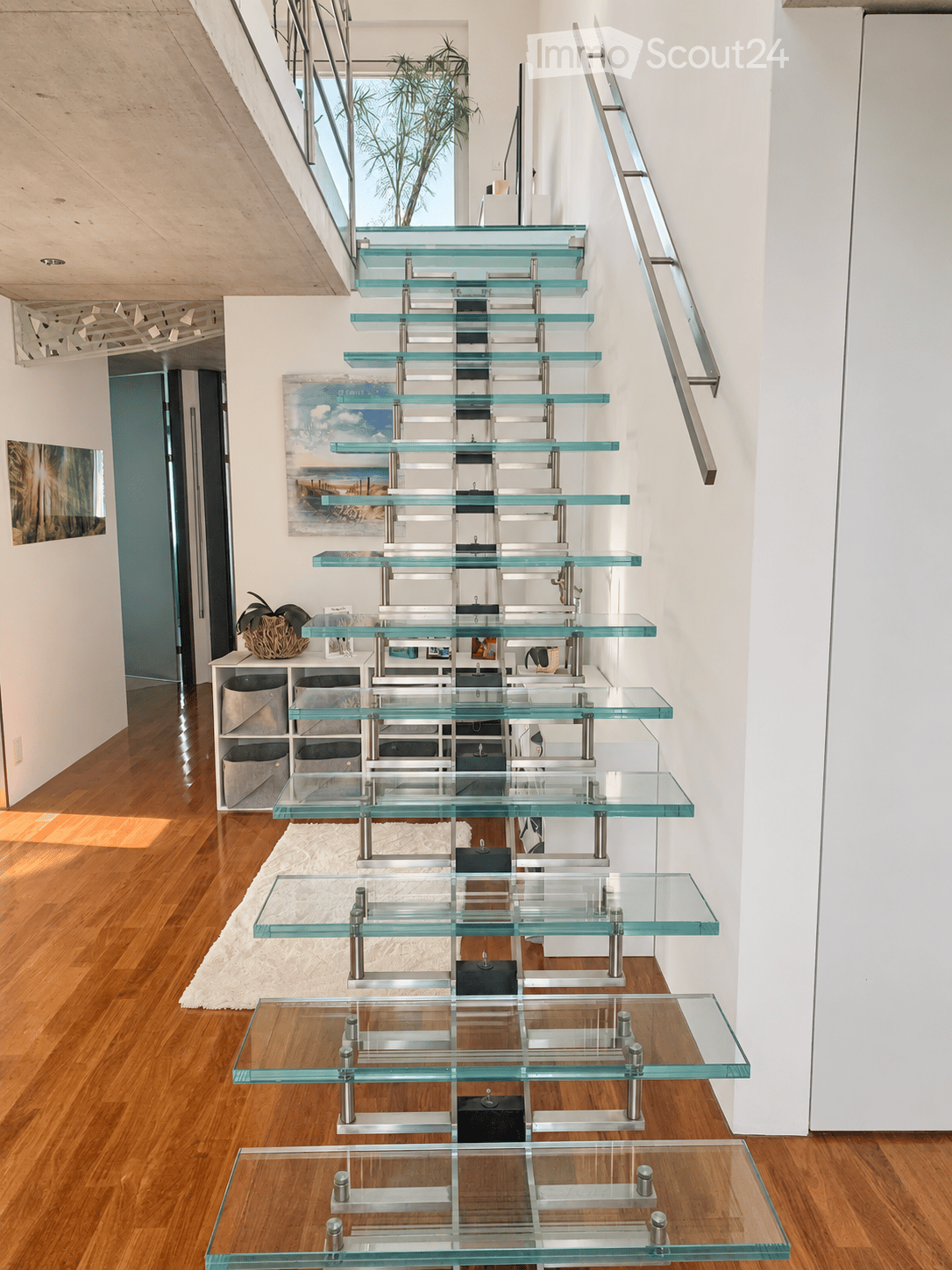 Modern hallway with a sleek glass staircase featuring metal railings, wooden flooring, and contemporary decor including a small shelf unit and artwork.