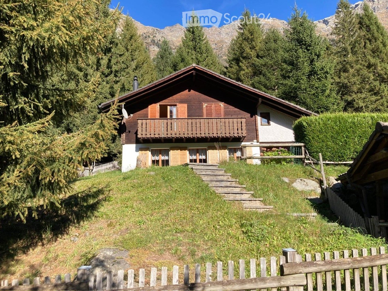Two-story wooden house with a balcony, surrounded by trees and mountains.