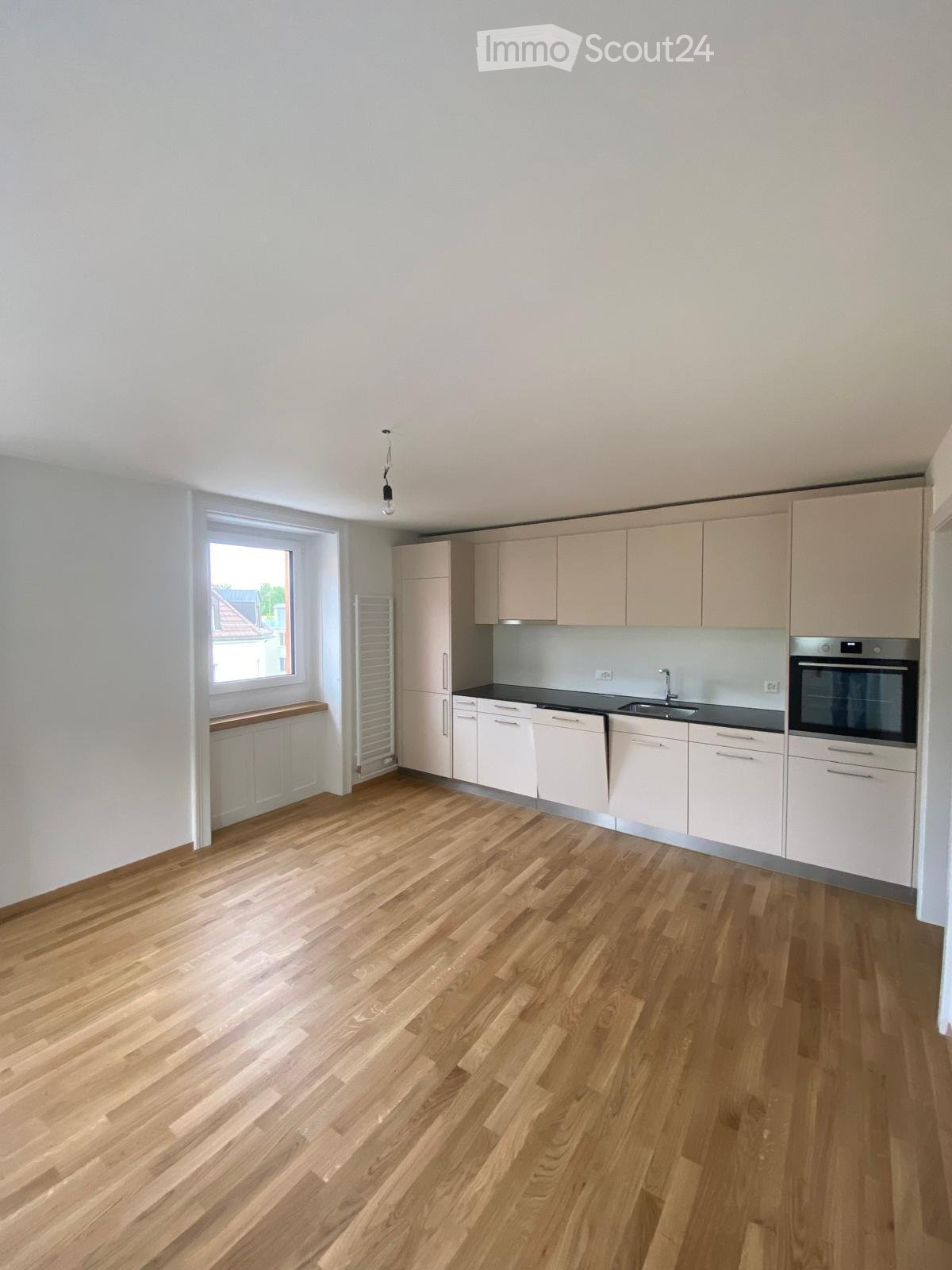 Empty kitchen with light wooden flooring, white cabinets, and a window letting in natural light. No appliances or furniture are present.