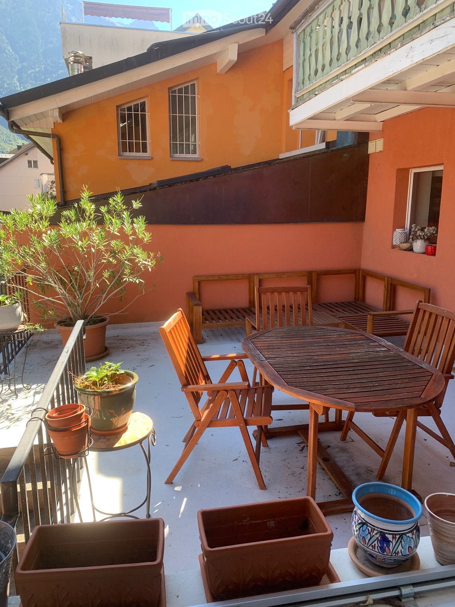 Balcony with wooden table, chairs, bench, and potted plants, overlooking a scenic mountain view.