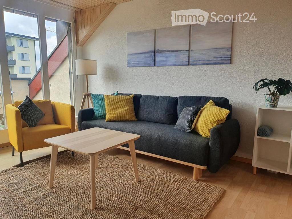 Modern living room with large windows offering a view, featuring a grey sofa with colorful pillows, a yellow armchair, a wooden coffee table, a floor lamp, a shelf with a plant, and abstract artwork on the wall.