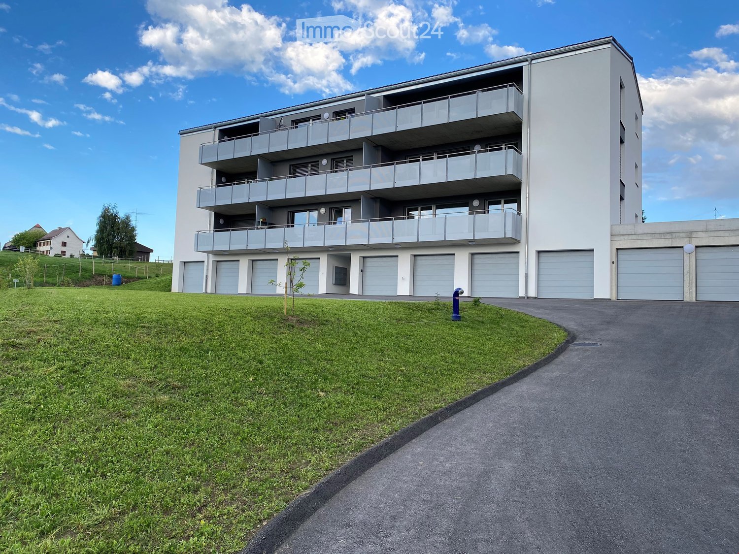 Modern multi-story apartment building with balconies and multiple garage doors, surrounded by a grassy area and a paved driveway.