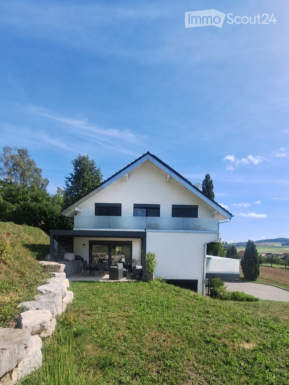 Modern two-story house with white exterior, black roof, and large windows. Features a raised ground floor, outdoor seating area, and landscaped garden.