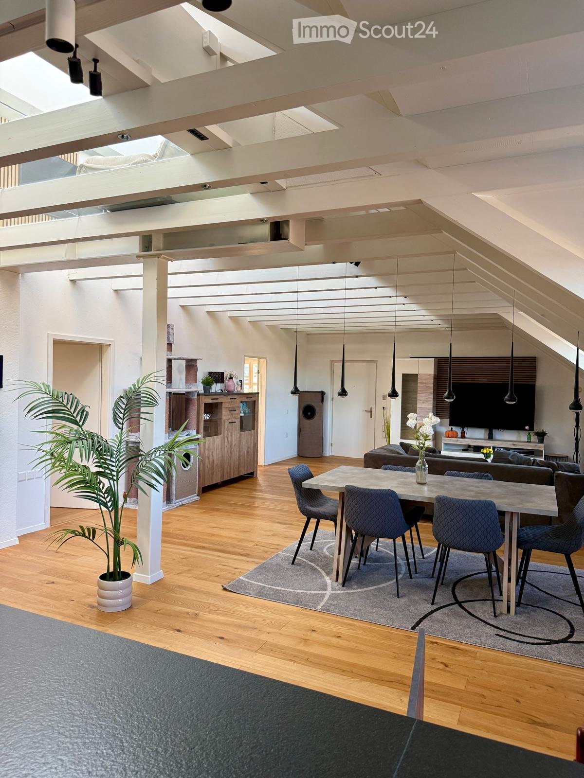 Open-concept living and dining area with wooden flooring, exposed wooden beams, pendant lights, a grey sofa, a dining table with four chairs, a cabinet, and a television on a stand.