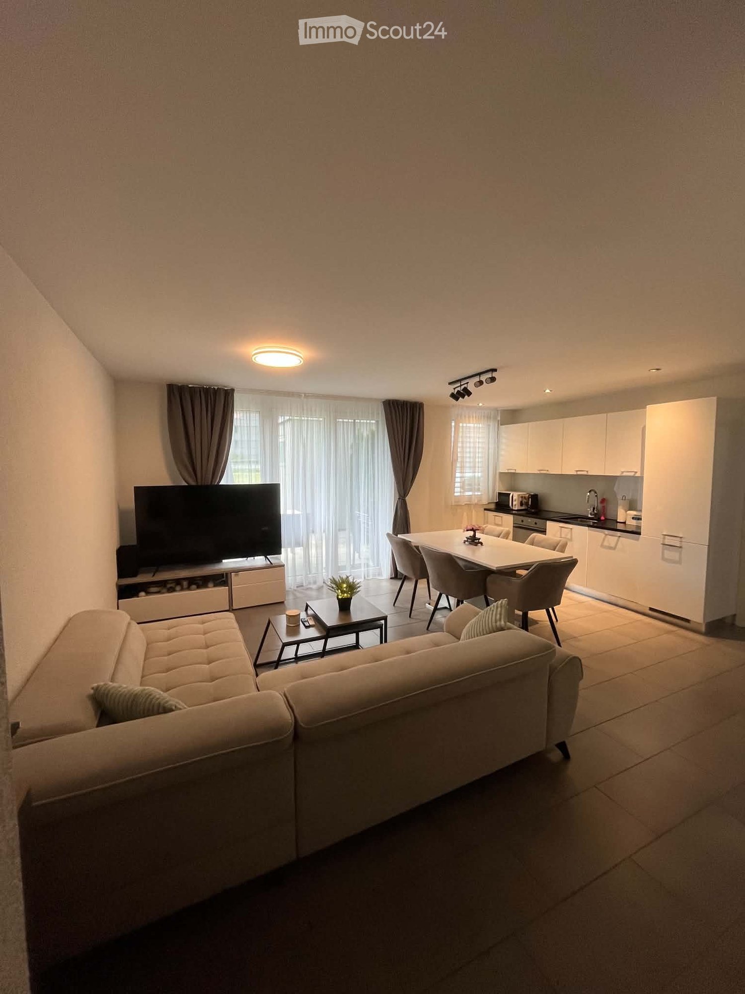 A modern, open-concept living and dining area with a light-colored sofa, coffee table, flat-screen TV, dining table with chairs, and a small kitchen area with white cabinets and a microwave.