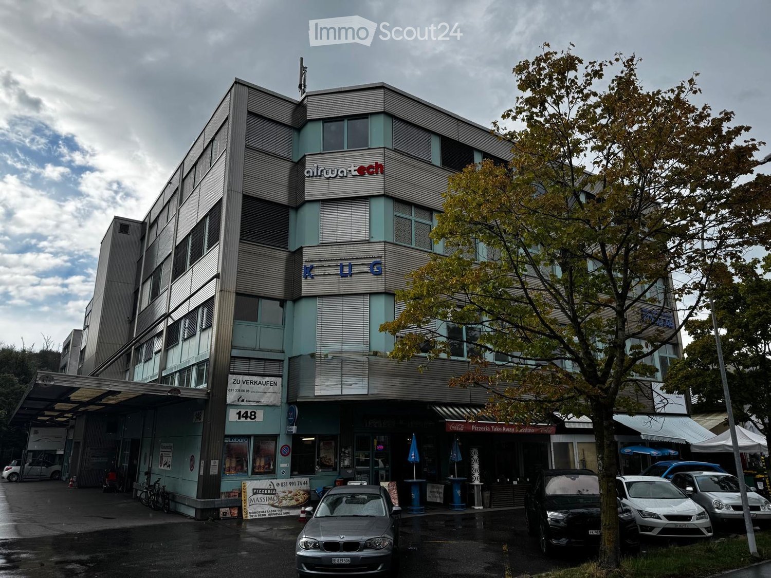 Modern multi-story building with signage for "airwatechnology" and "KUG" on the facade, with a pizza take-away storefront and several parked cars in front.