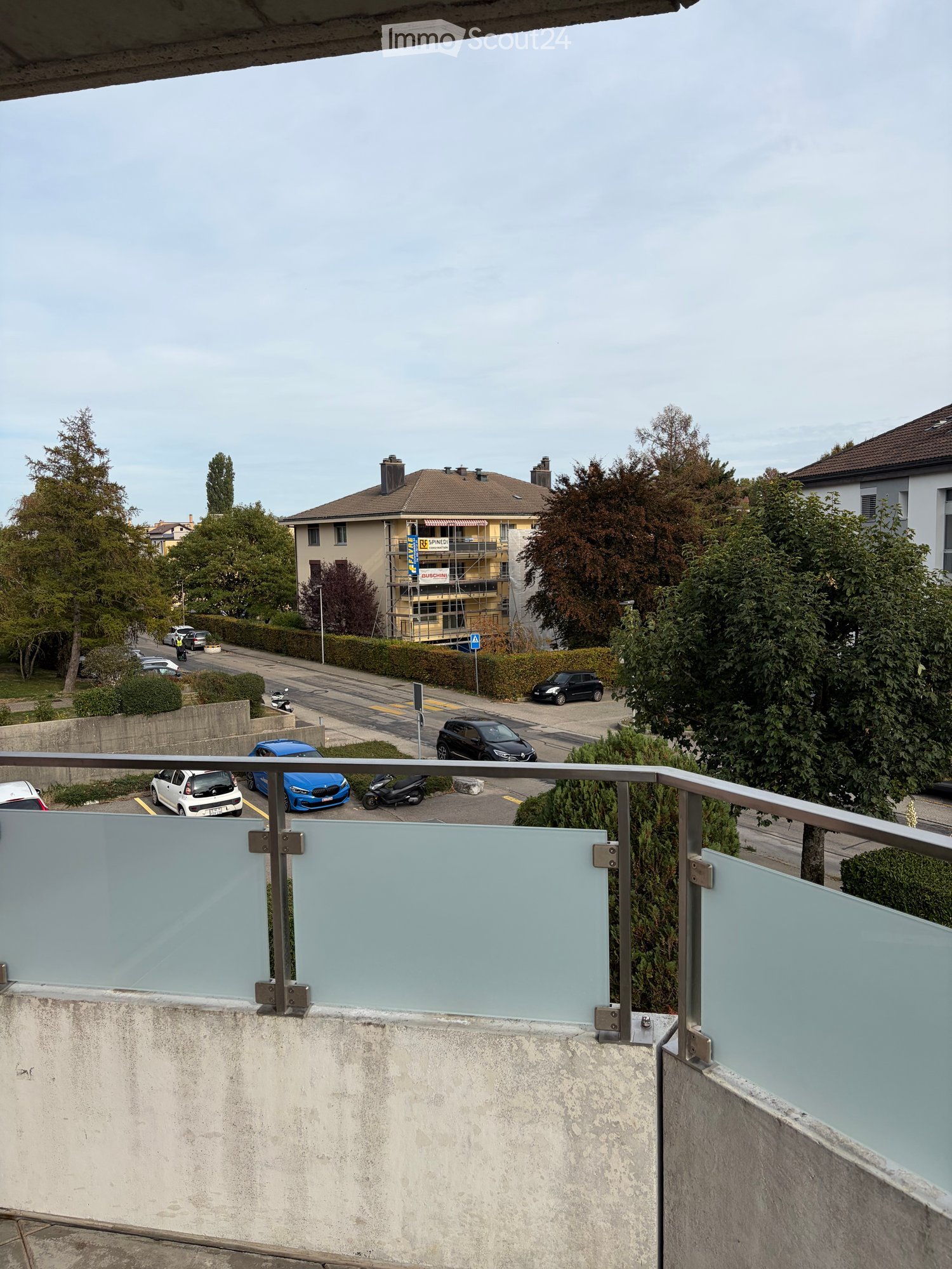 Balcony with glass railing overlooking a street with parked cars and buildings in the background.