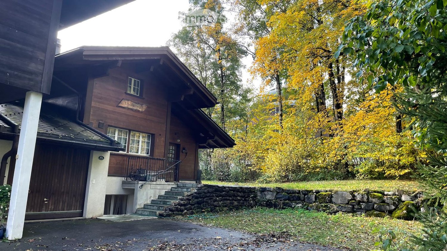 A two-story wooden house with a stone wall and steps leading to the entrance, surrounded by autumn foliage and trees.