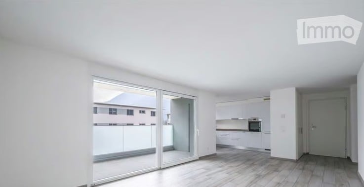 Bright, empty room with large sliding glass doors leading to a balcony, white walls, light wooden flooring, and a small adjacent kitchen area with white cabinets and appliances.