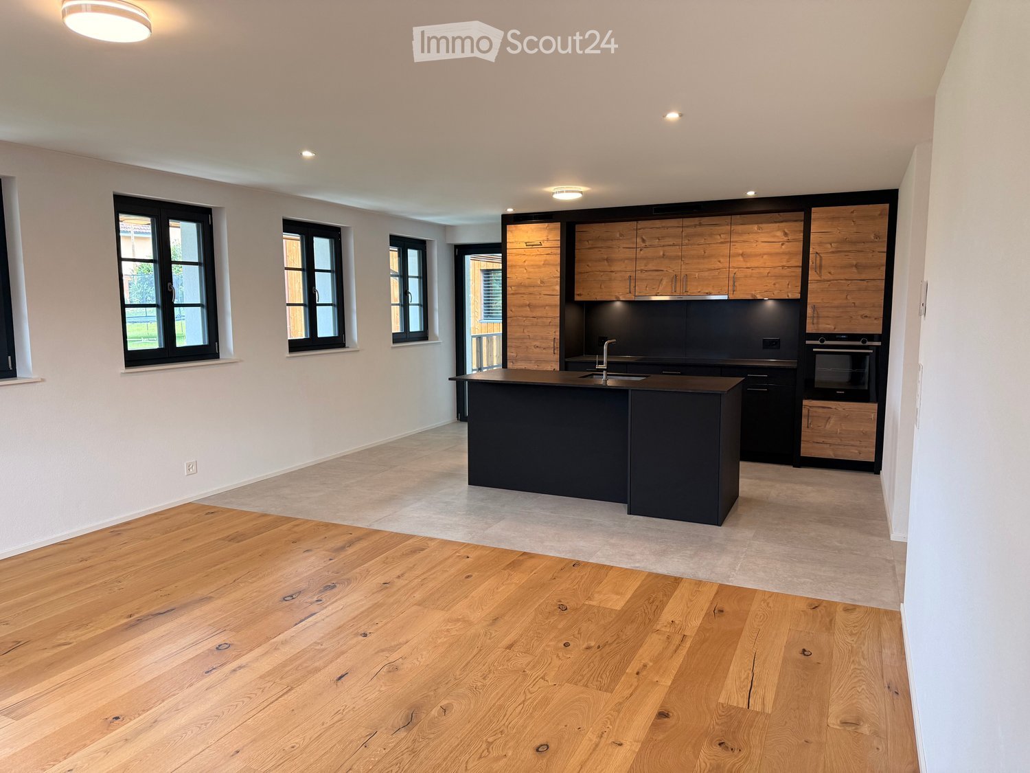 Modern kitchen with wooden cabinetry, black island, and stainless steel appliances. Large windows provide natural light.