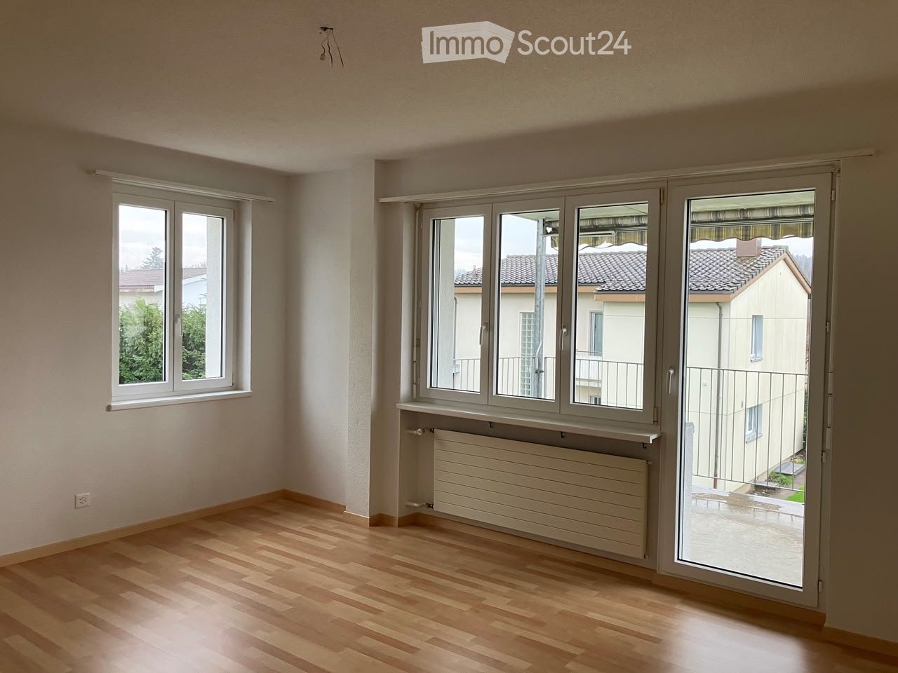 Empty room with wooden flooring, large windows, radiator, and a sliding glass door leading to a balcony.