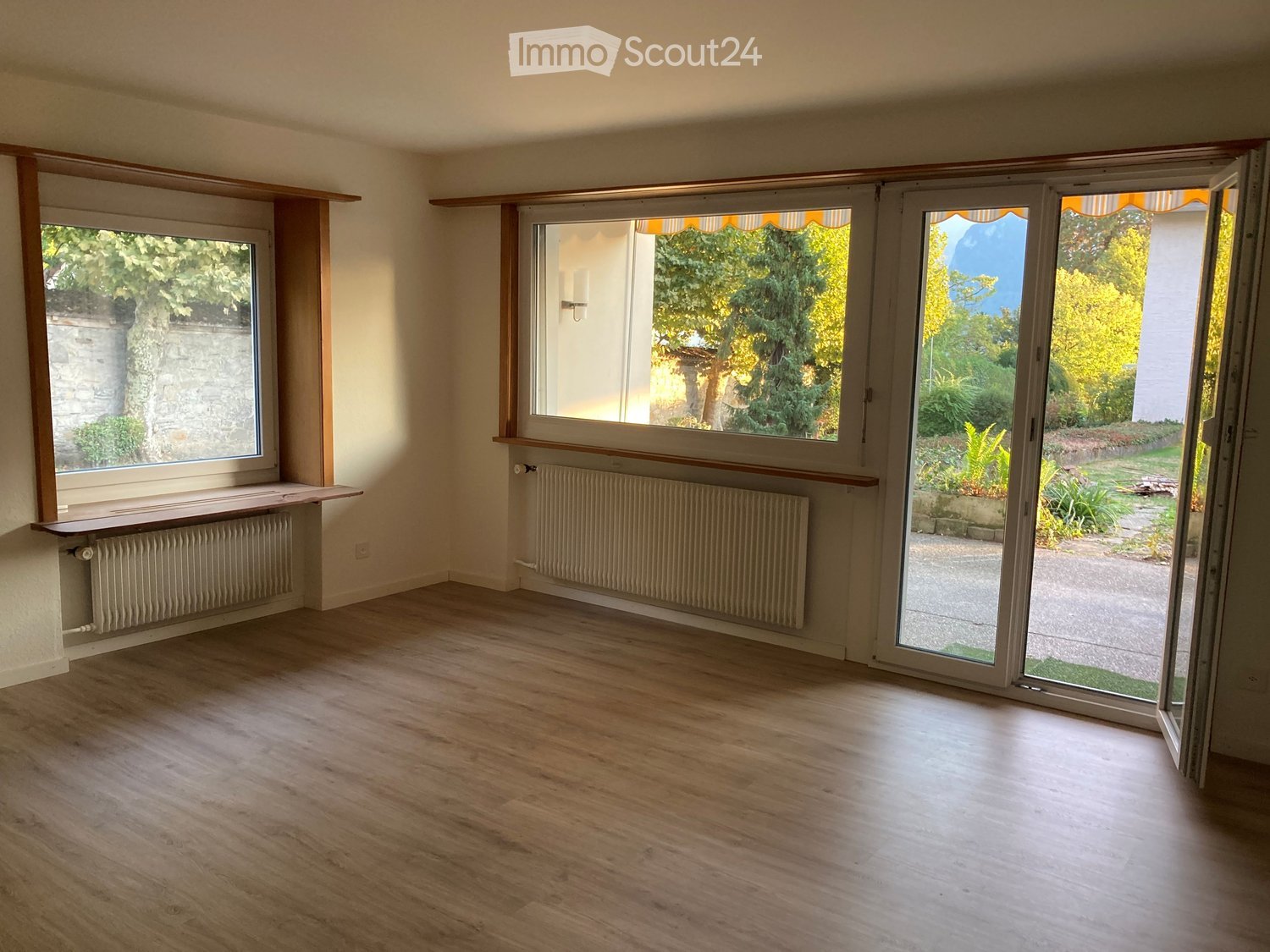 Empty room with wooden flooring, large windows, radiators, and sliding glass doors opening to a garden.