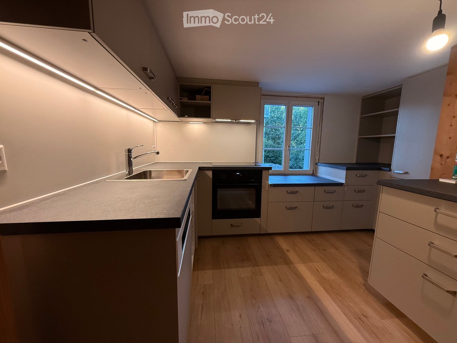 Modern kitchen with light-colored cabinets, stainless steel sink and faucet, built-in oven, and wooden flooring.