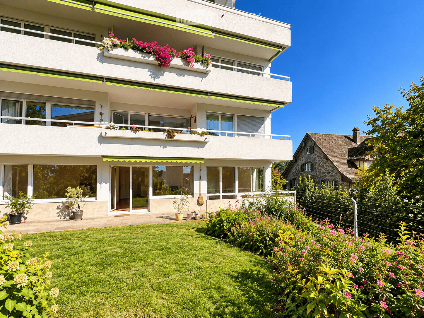 Modern three-story apartment building with balconies adorned with colorful flowers, large windows, and a well-maintained garden in front.