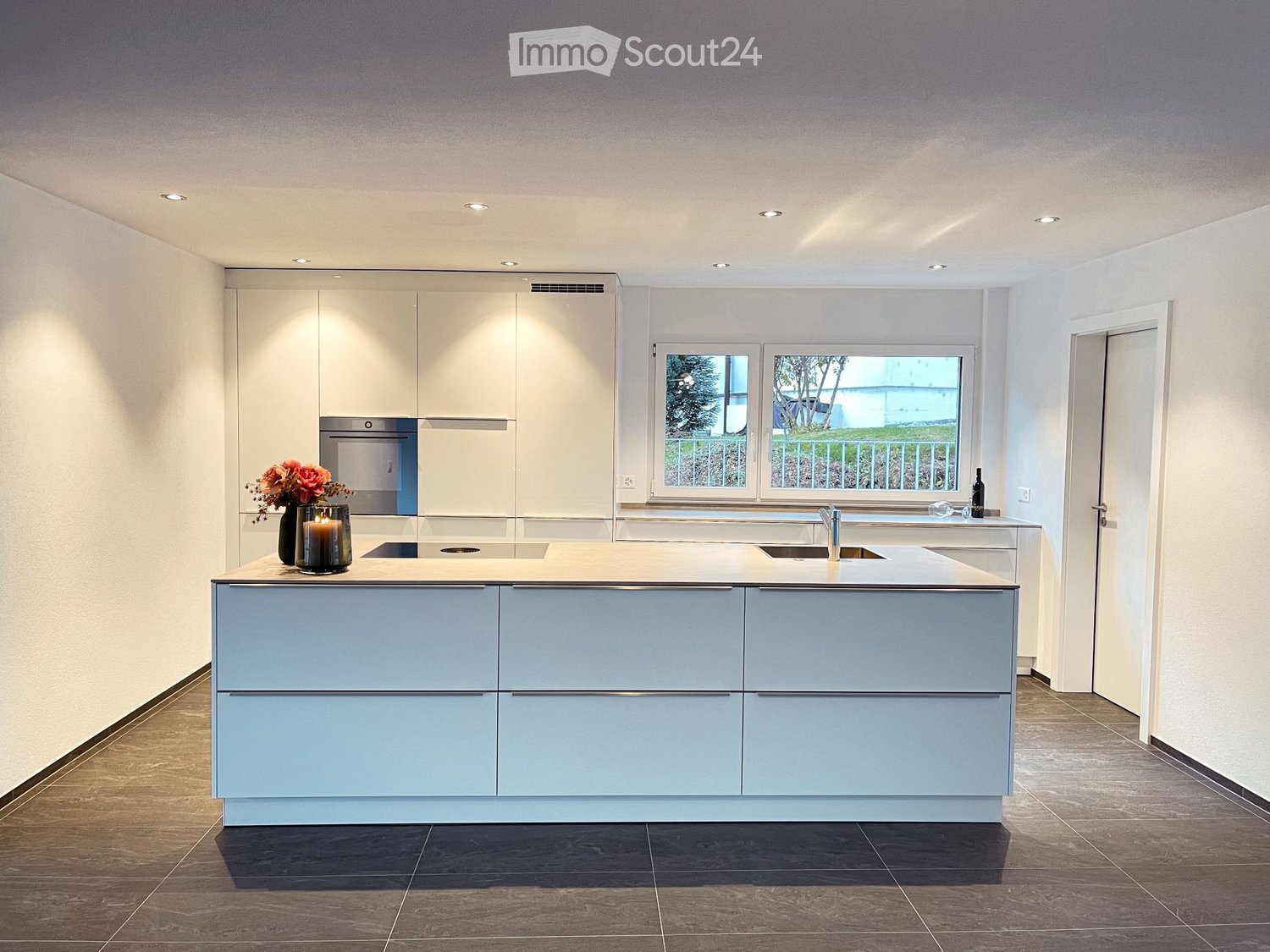 Modern kitchen with a central island, built-in cabinets, and a large window overlooking a green area. Features include a sink, faucet, candle, vase with flowers, and a bottle on the counter.