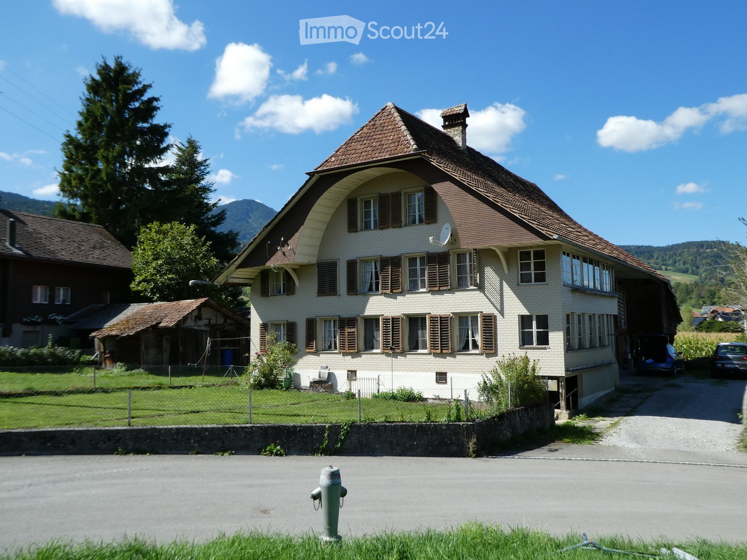 Two-story house with a brown tiled roof, multiple windows with shutters, and a satellite dish on the roof. The house is surrounded by a low stone wall and a grassy area with trees and mountains in the background.