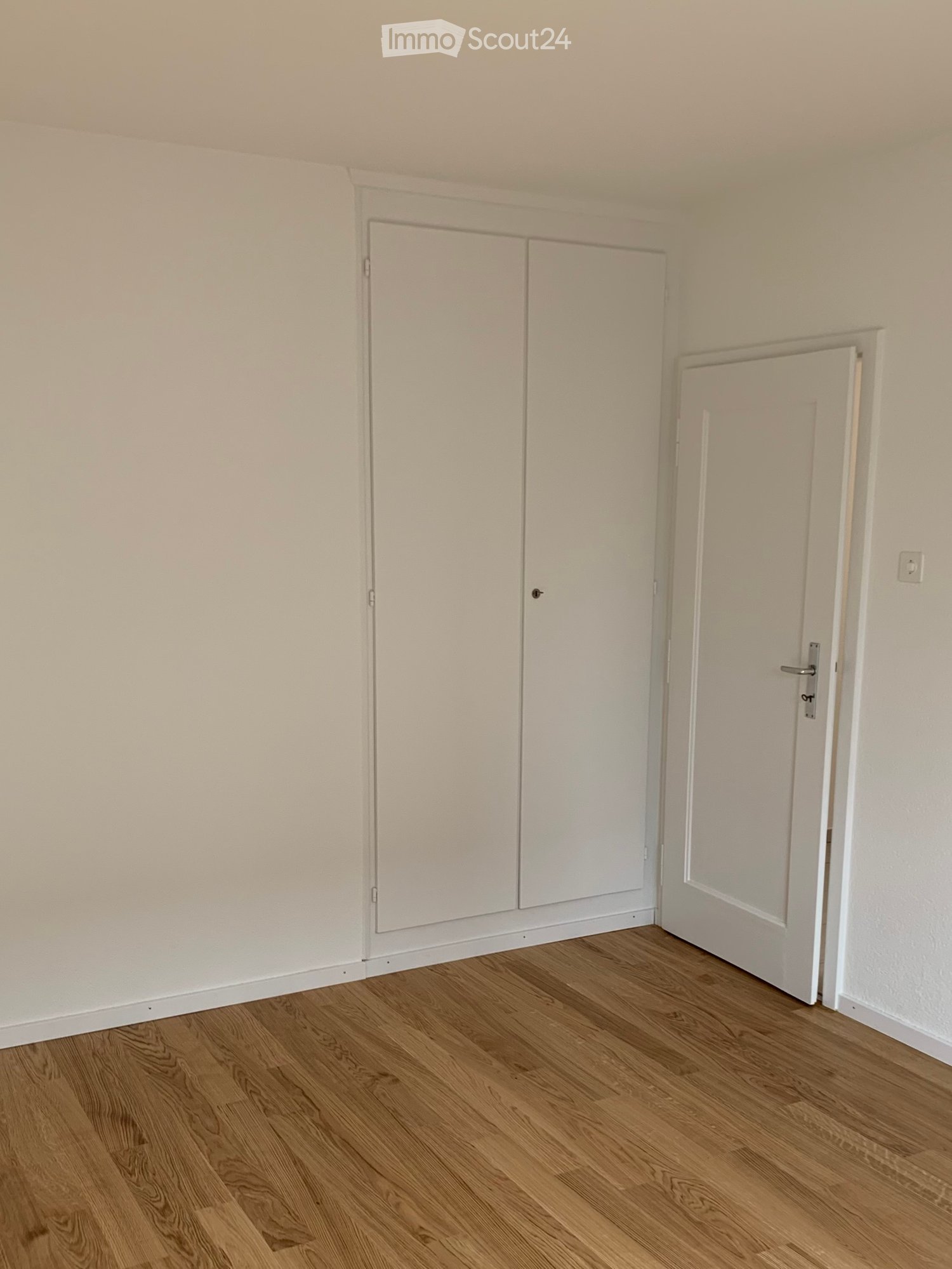 Empty room with light-colored walls and wooden flooring, featuring a closed double-door closet and an open doorway leading to another space.