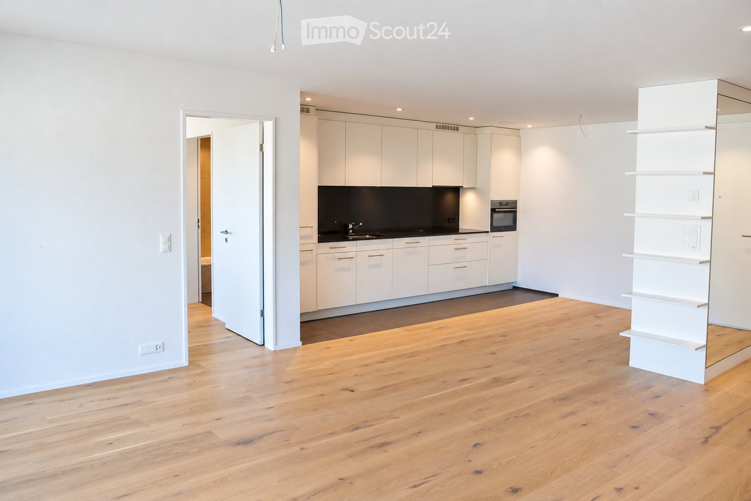 Open-plan living and kitchen area with light wooden flooring, white kitchen cabinets, a sink, and an oven.