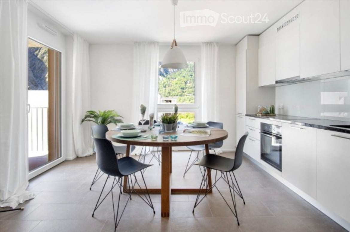 Open-plan kitchen and dining area with white cabinets, a round wooden dining table, modern chairs, and a pendant light. Large windows offer a scenic view.