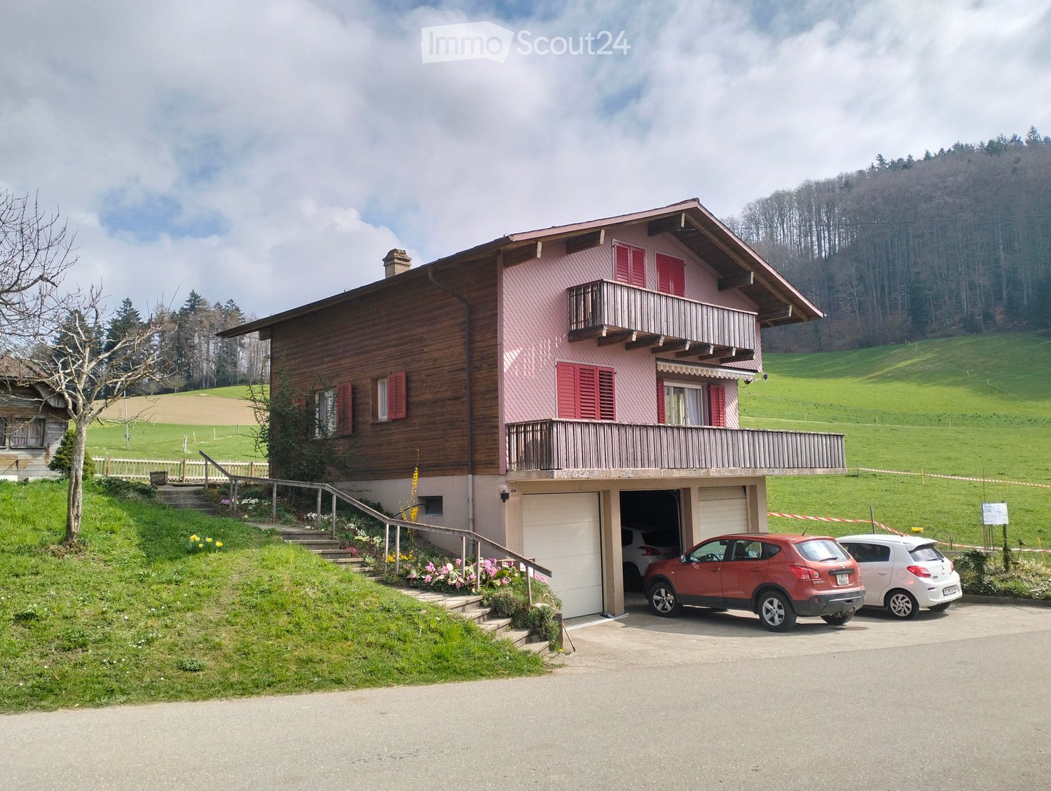 Two-story house with pink siding, red shutters, balcony, garage, and two parked cars.