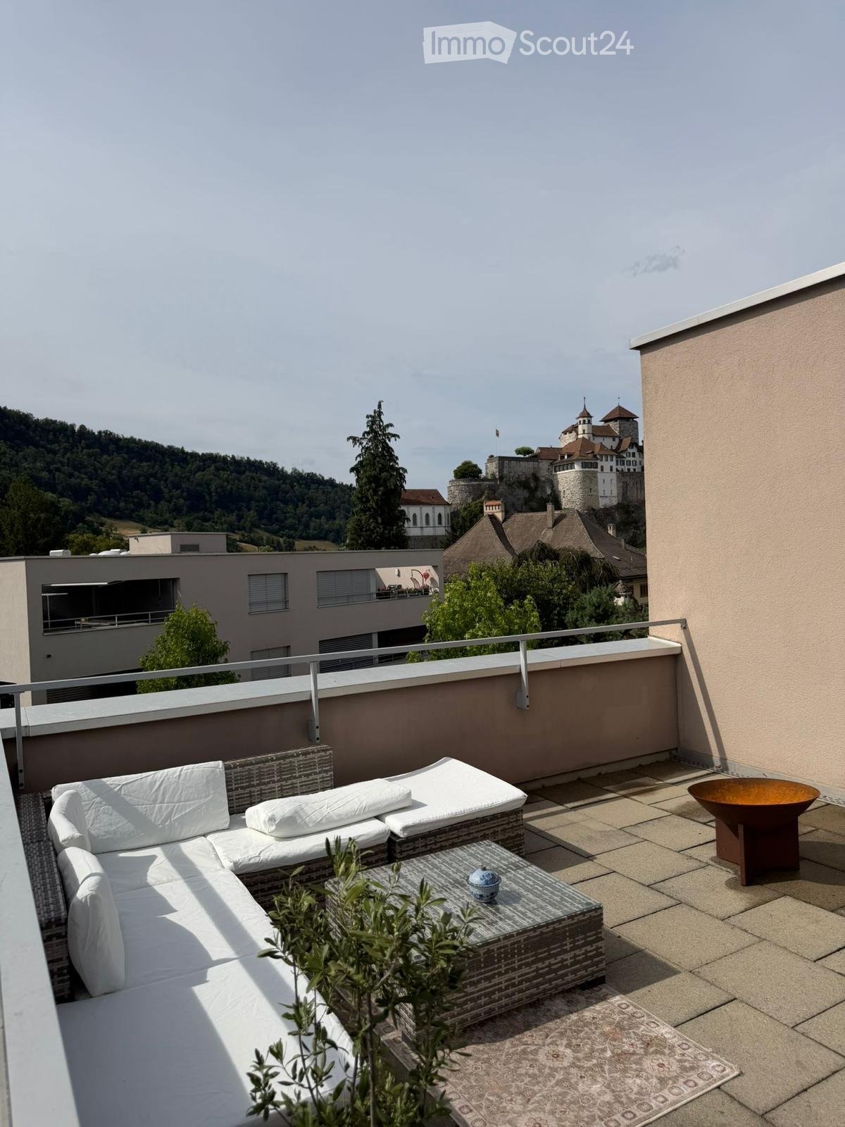 Rooftop terrace with white sofa, coffee table, small table with a bowl, and a decorative fire pit, overlooking a scenic town and castle.