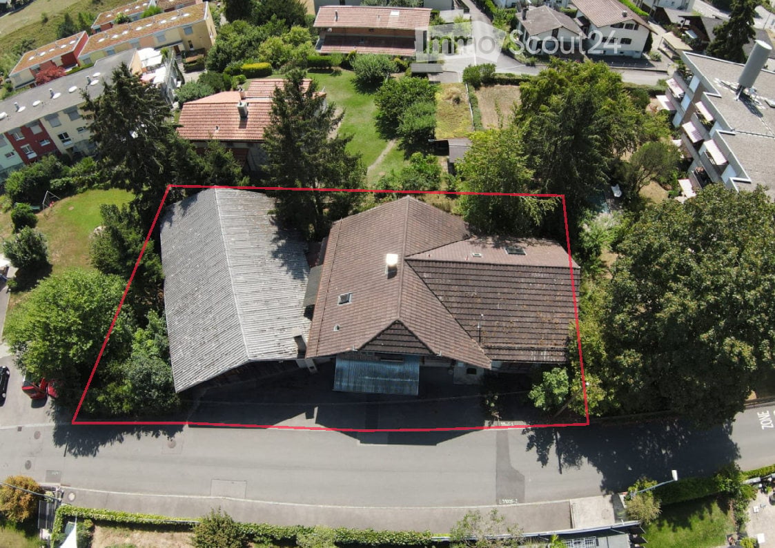 Aerial view of a two-story house with a gray roof and multiple chimneys, surrounded by trees and neighboring houses.